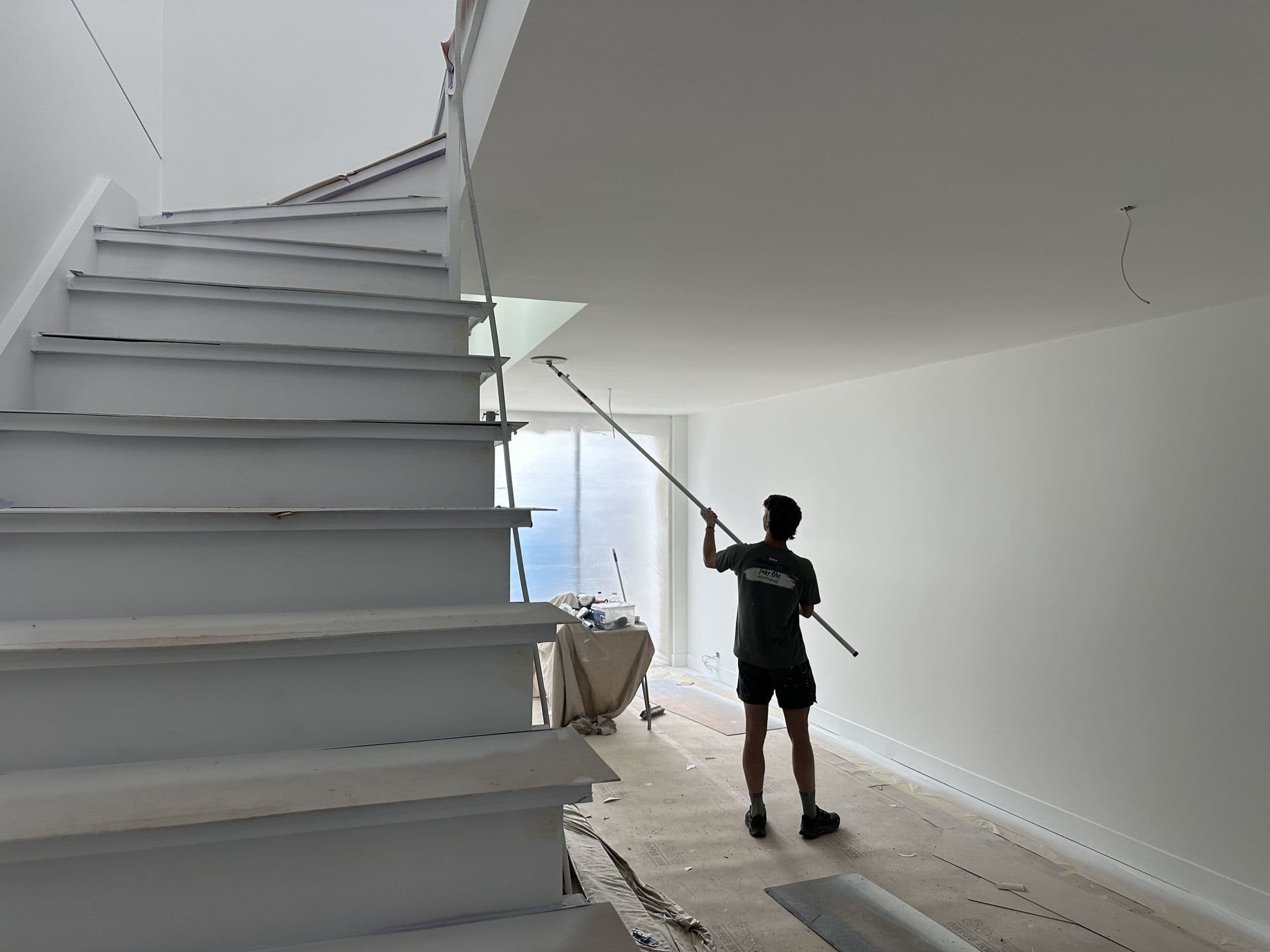 Tricoat painter cutting in a wall beside a sculptural glass-balustrade staircase, mid-job